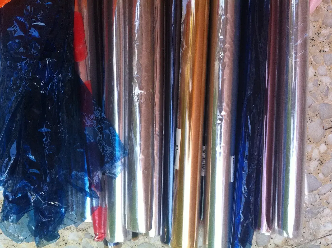 Tightly packed rolls of cellophane film in a rainbow of colors (blue, red, silver, gold, and pink) stacked upright and catching the light.