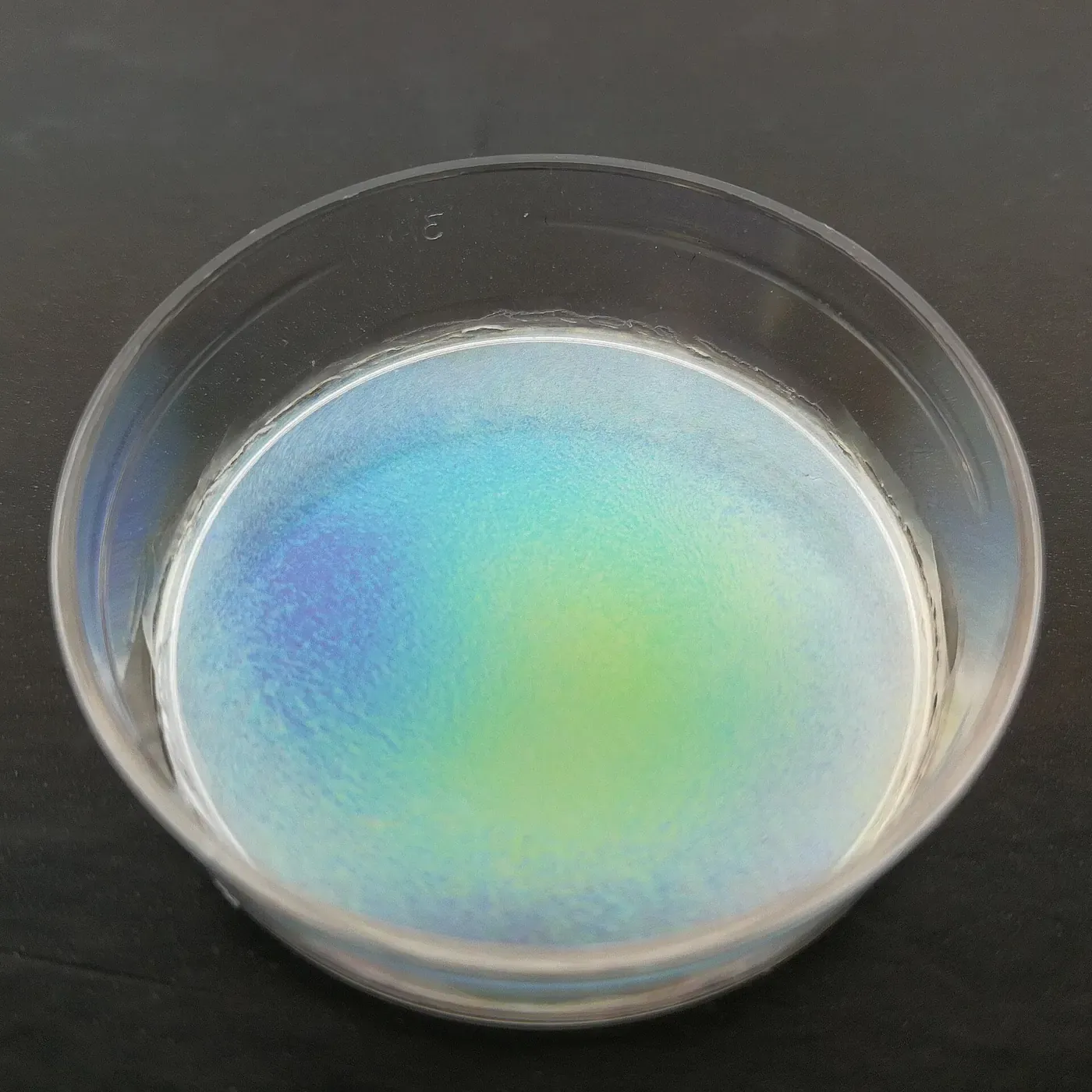 A shallow glass petri dish containing a thin, dried film that shimmers in bands of blue, green and purple, resting on a dark surface.