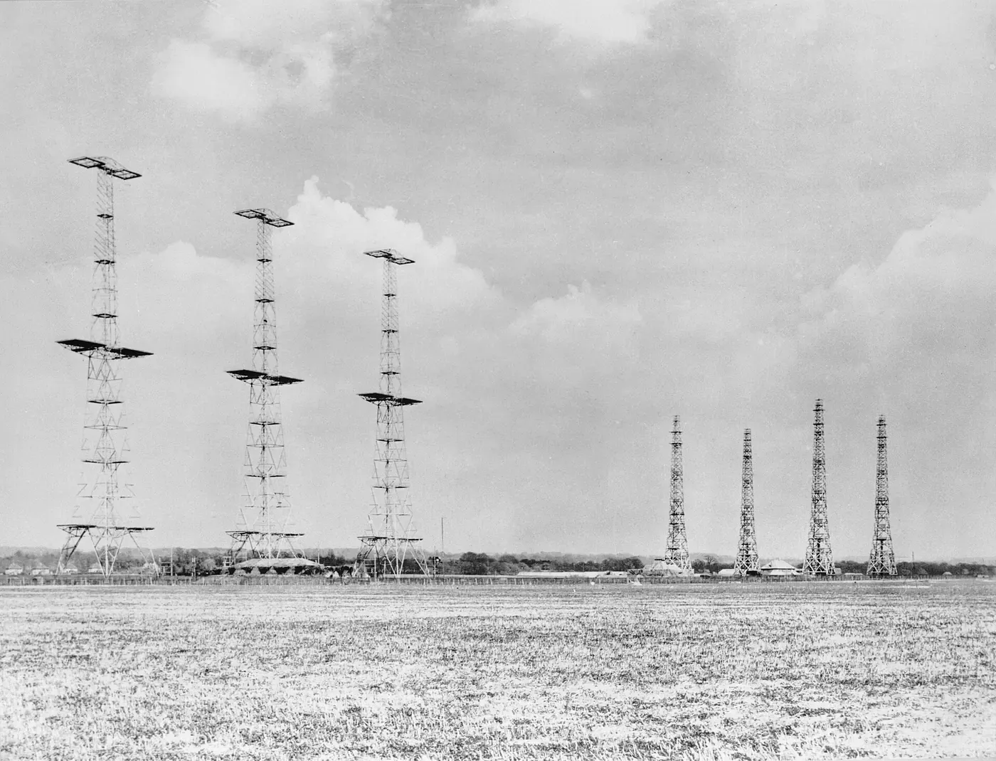 Seven tall steel and timber lattice radar towers standing in a flat field, photographed from ground level against a cloudy sky.