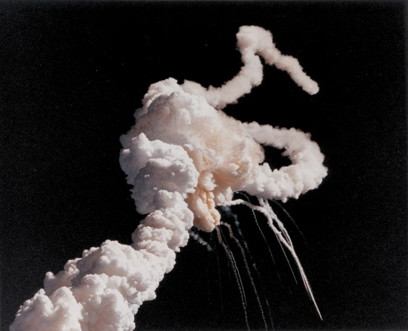 A billowing white and orange cloud of exhaust and debris against black sky, with thin smoke trails streaking downward from it, a few seconds after an in-flight breakup.