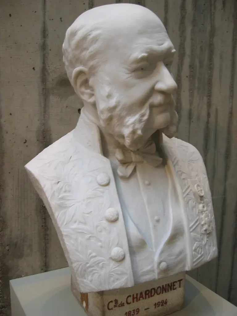 A white marble bust of a balding, bearded man in formal dress with a bow tie and a decoration on his lapel, on a plinth reading 'Cte de Chardonnet, 1839-1924.'