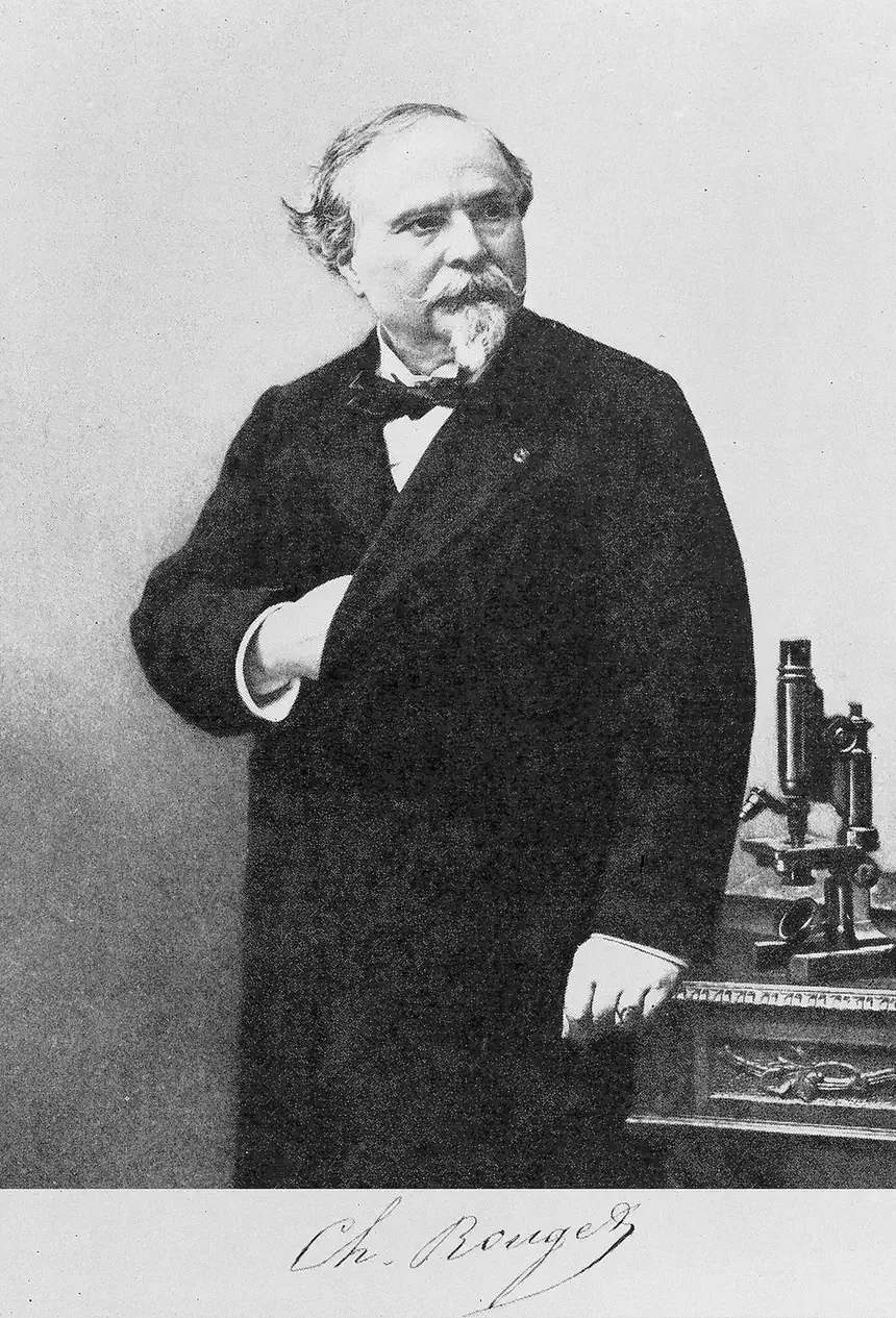 A formal 19th-century studio portrait of a moustached man in a dark coat and bow tie, standing beside a brass microscope, with his signature printed below the image.