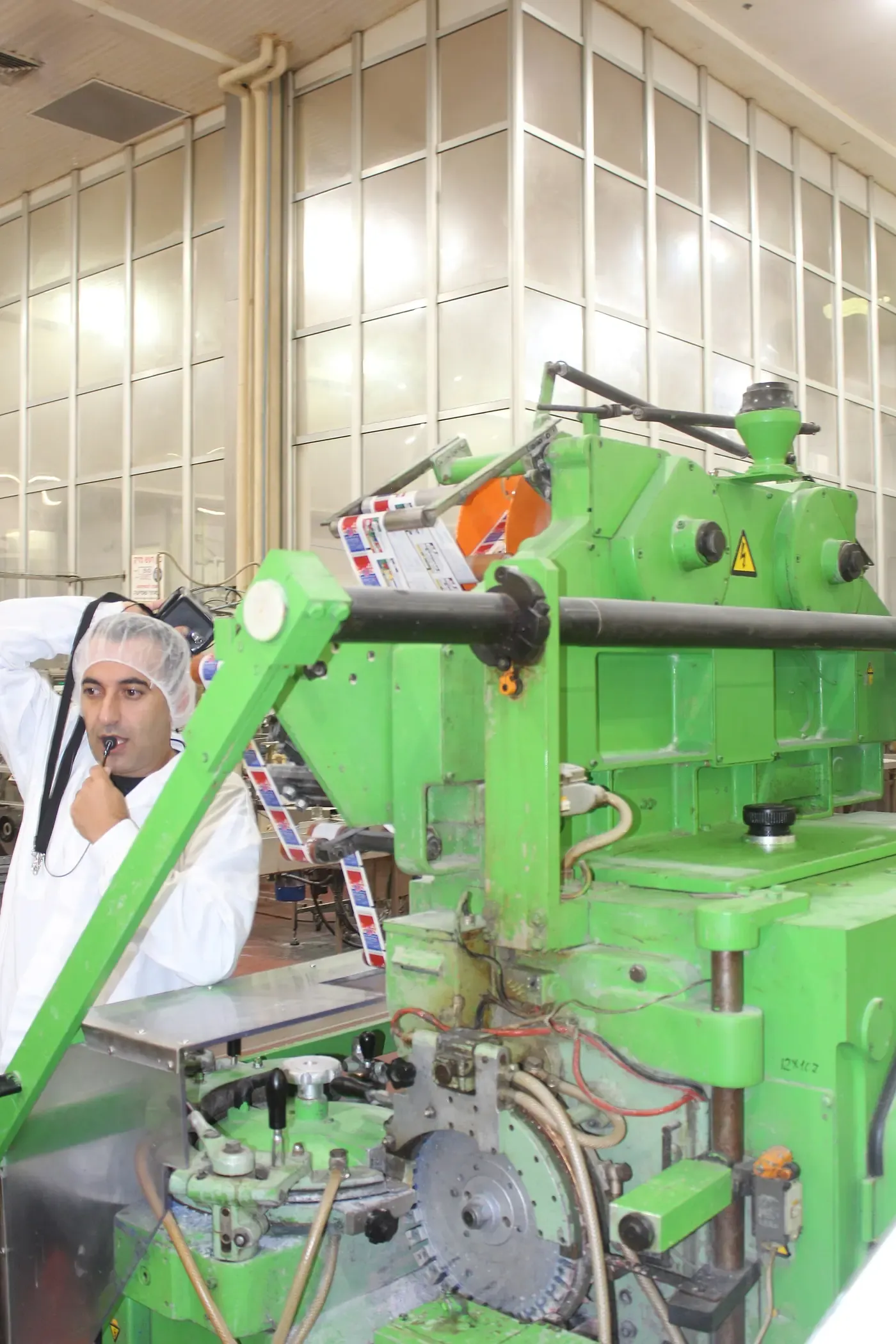 A factory worker in a hairnet and white coat standing beside a large green industrial machine that folds and wraps strips of gum on a production line.