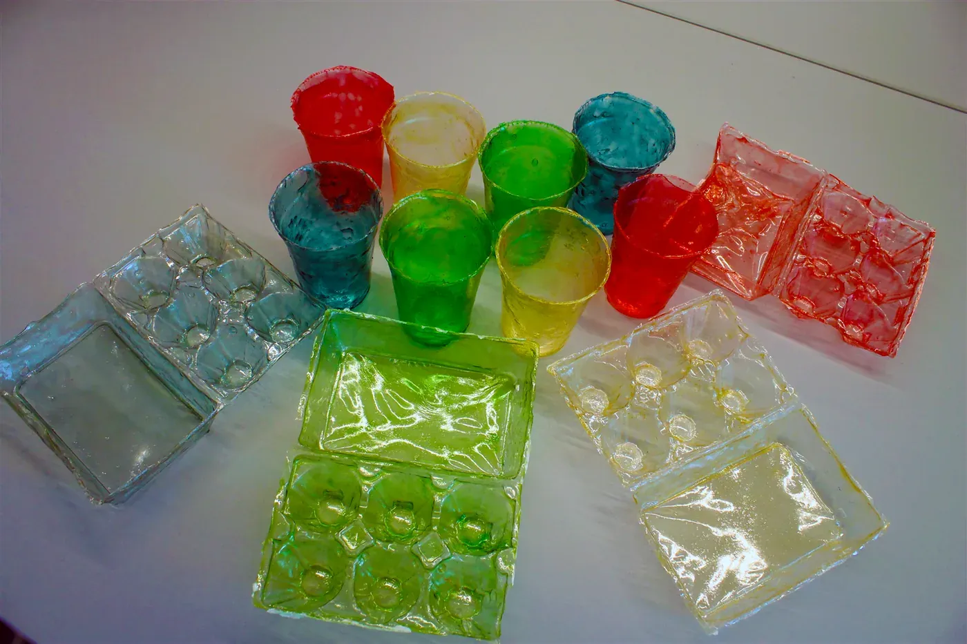 An arrangement of small brightly coloured translucent cups and moulded trays with egg-carton-style indentations, in red, yellow, green and blue, laid out on a white tabletop.