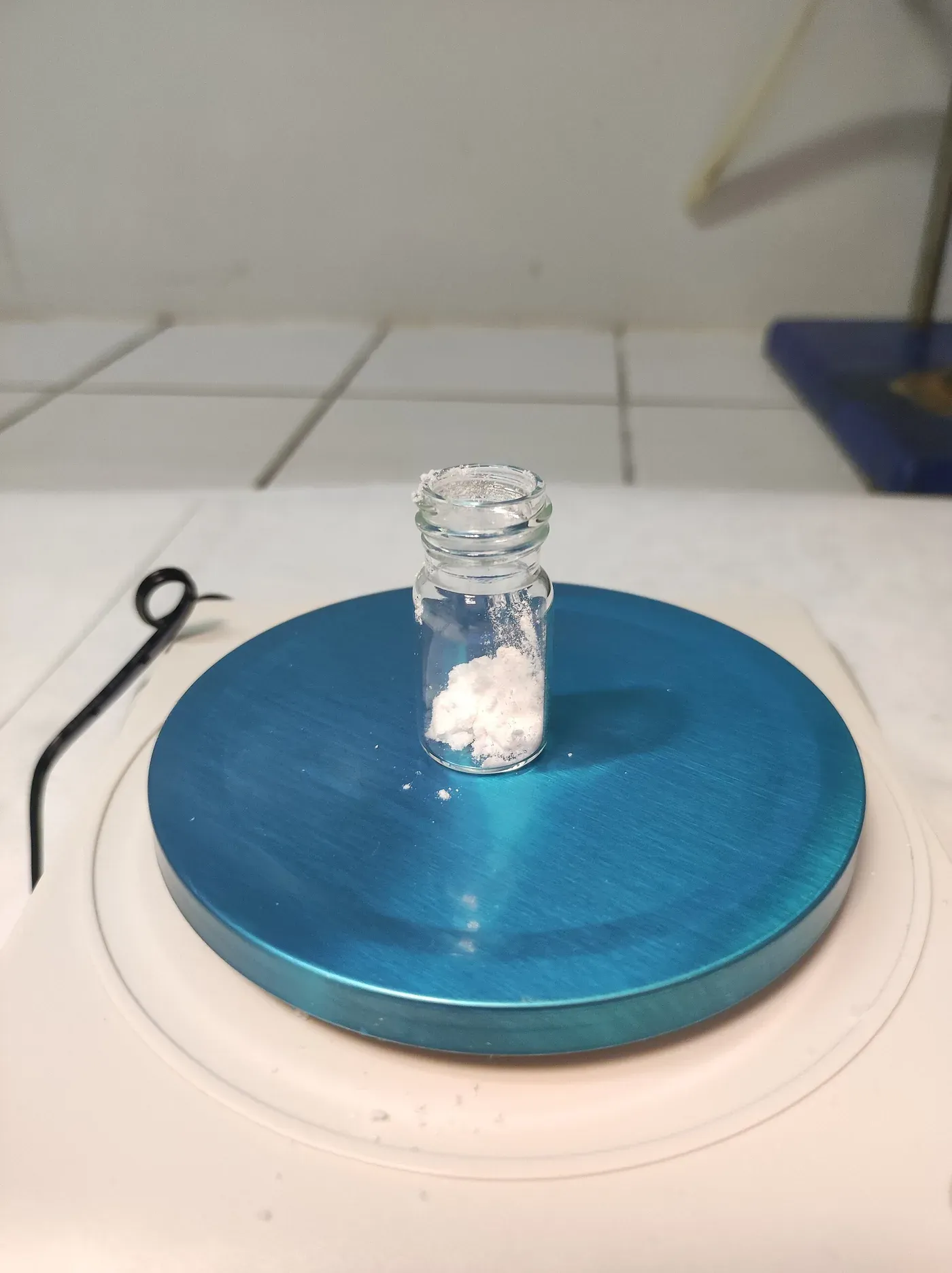 A small open glass vial containing a clump of fine white powder, standing on a blue laboratory hotplate stirrer, with an analytical balance visible in the background.