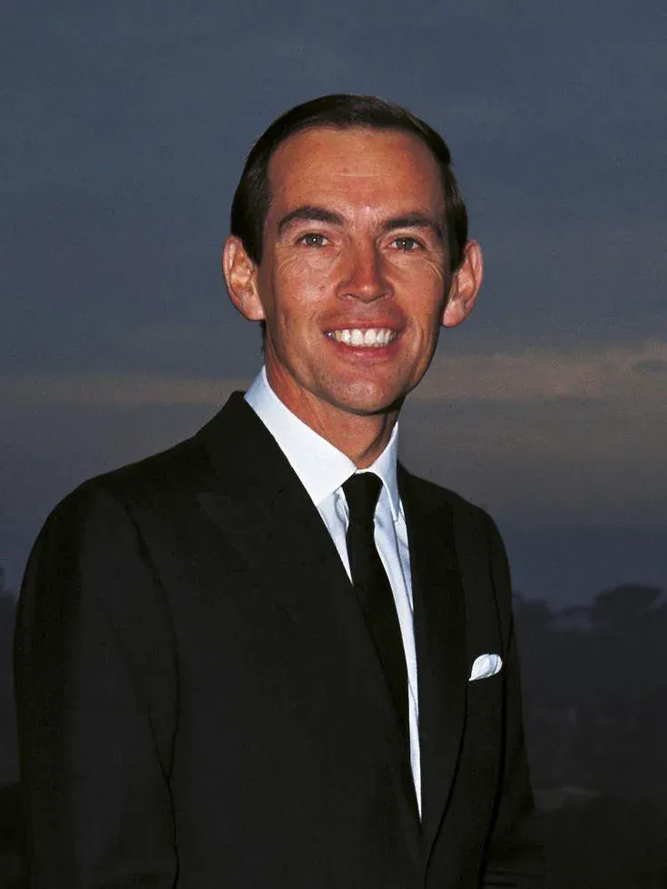 A head-and-shoulders portrait of a smiling man with dark hair, wearing a black suit and tie with a white pocket square, photographed outdoors at dusk.