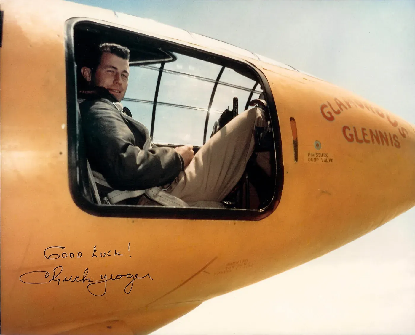 A close-up photograph looking into the open cockpit window of the bright orange Bell X-1 rocket plane, with pilot Chuck Yeager visible inside in flight gear; the aircraft is marked 'Glamorous Glennis', and a later autograph reading 'Good Luck! Chuck Yeager' is written across the lower part of the photograph.
