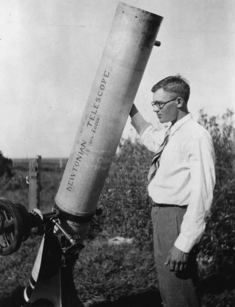 A young man in glasses, shirt and tie standing beside a tall homemade telescope tube mounted on an equatorial stand in an open field.
