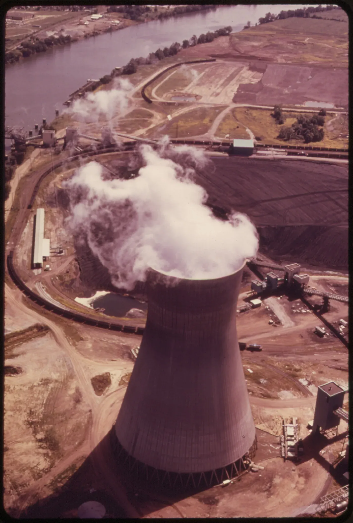 An aerial view of a coal-fired power plant's cooling tower venting a large plume of steam, with coal stockpiles, rail lines and a river visible in the surrounding landscape.