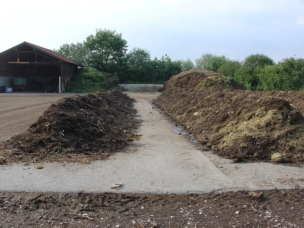 Two long windrows of dark, decomposing organic material piled beside a paved yard, with an open-sided barn and trees in the background.