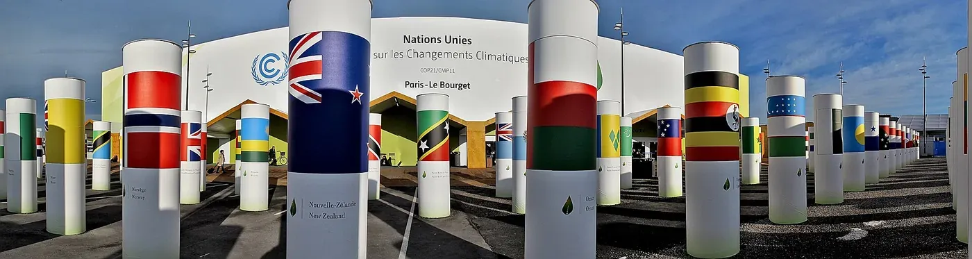 A row of tall white columns painted with national flags and country names, forming an avenue leading toward a large white conference building marked 'Nations Unies sur les Changements Climatiques, COP21/CMP11, Paris–Le Bourget'.
