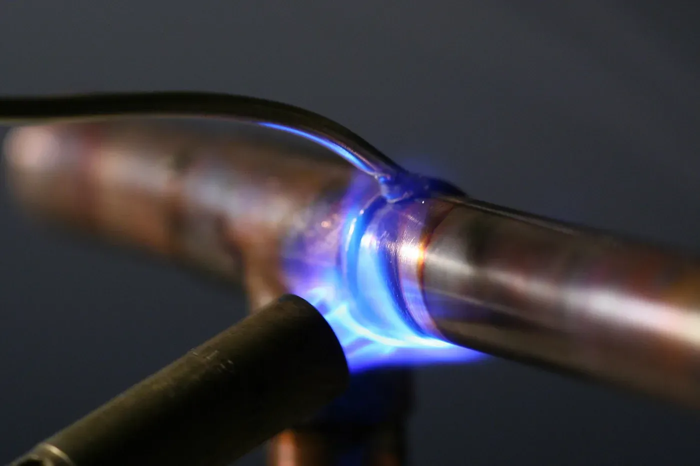 A close-up of a blue propane-torch flame heating a joint between two copper pipes, with a bright bead of melted solder running around the fitting.