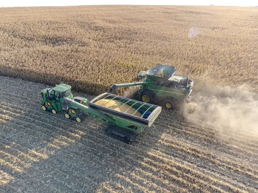 An aerial photograph of a green combine harvester cutting through a large field of dried corn stalks, unloading grain into a cart towed alongside it, with dust rising behind.