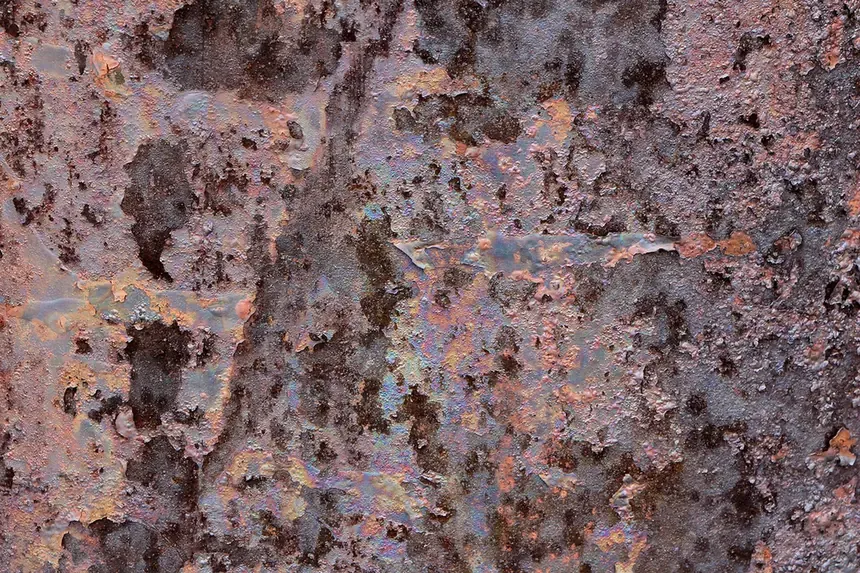 A close-up of a heavily corroded steel surface, its rust flaking away in mottled patches of orange and brown with an iridescent blue-purple sheen.