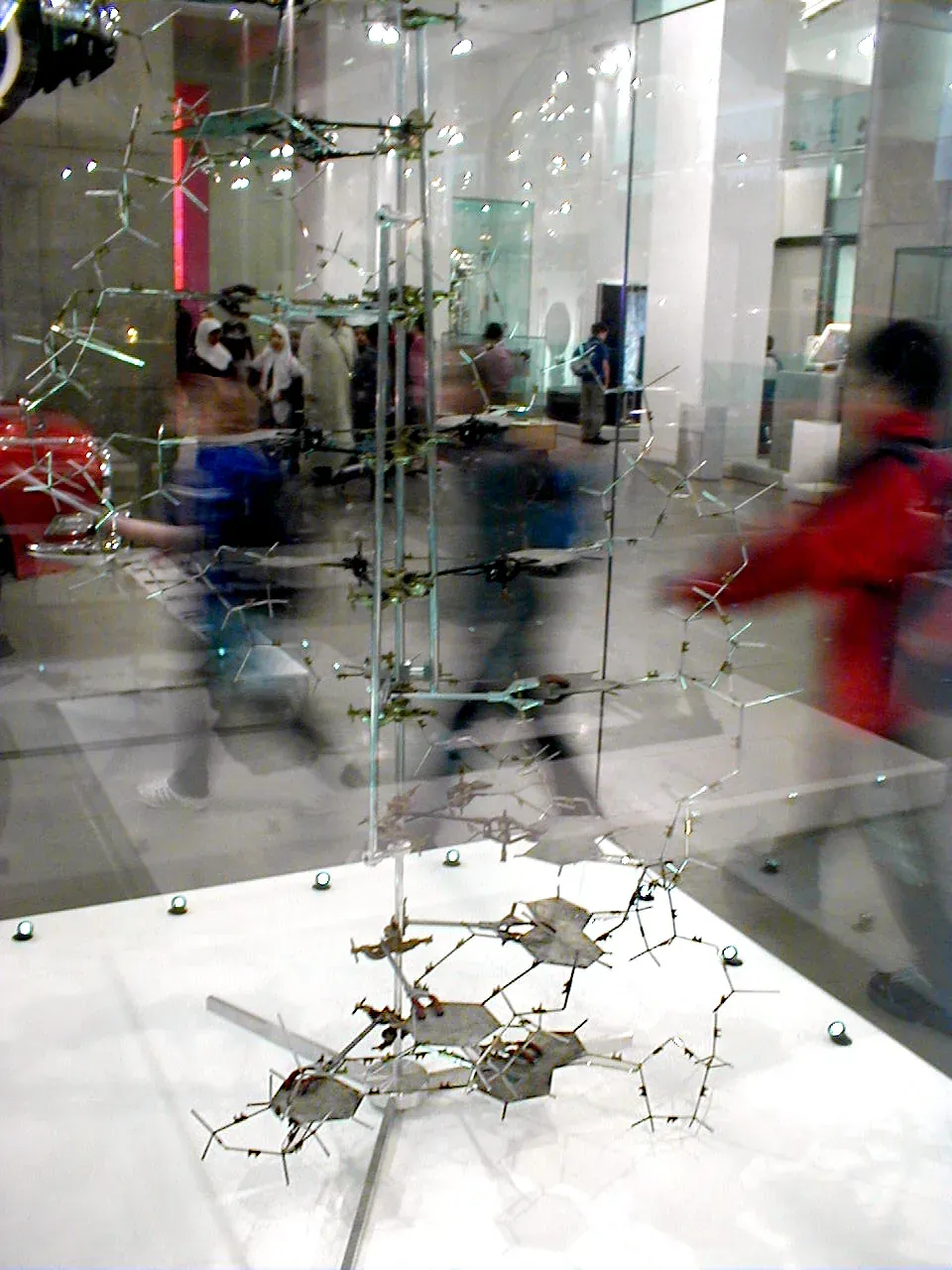 A tall double-helix-shaped model built from wire rods and flat metal plates, displayed in a museum glass case, with the blurred figures of visitors passing in front of it.