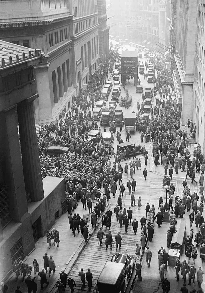 A high-angle photograph looking down a narrow street between tall stone office buildings, packed edge to edge with a crowd of people in coats and hats, automobiles stalled in the crush below.