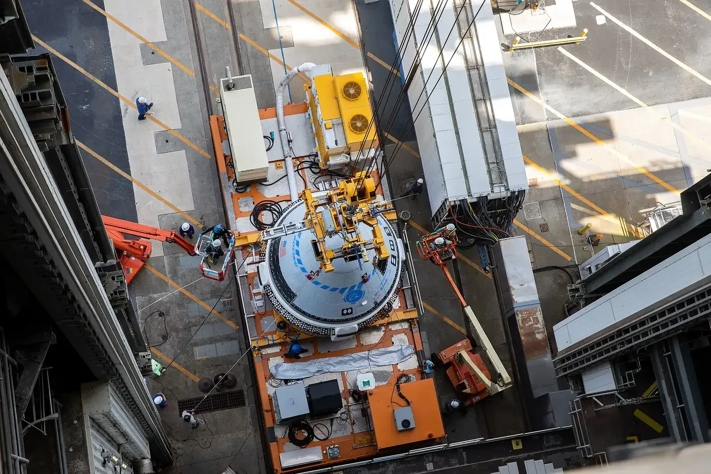 An overhead view of a white, dome-topped space capsule being lifted by an overhead crane inside an industrial processing building, surrounded by scaffolding, work platforms, and technicians in hard hats.
