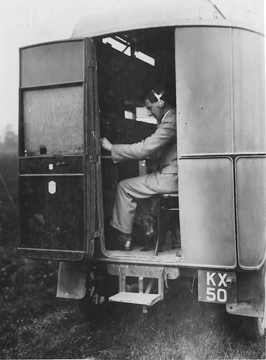 A man wearing headphones sits inside the open rear doors of a boxy 1930s radio van, operating receiver equipment, with a small dog at his feet.