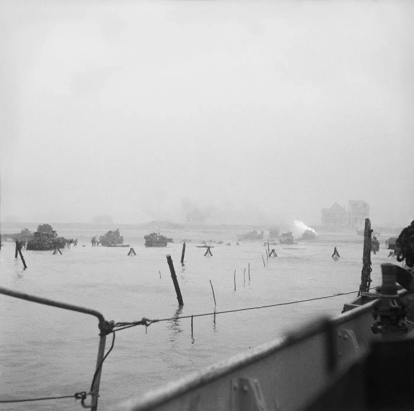 A hazy black-and-white photograph, taken from a landing craft, of tanks and vehicles disembarking onto a beach among rows of wooden anti-invasion stakes driven into the shallows.