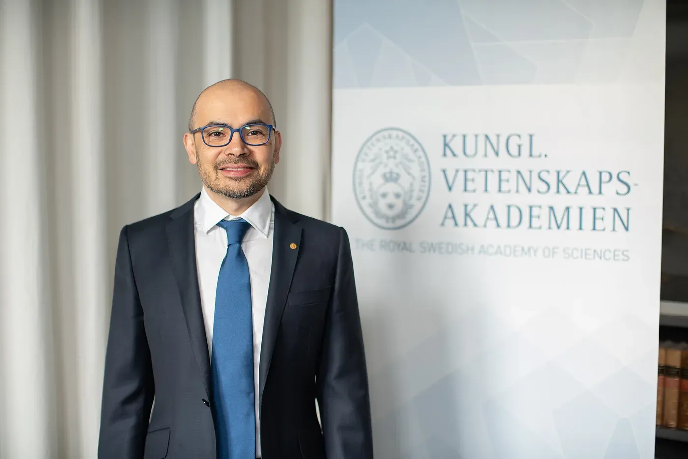 A smiling bald man wearing glasses and a dark suit with a blue tie stands beside a banner reading 'Kungl. Vetenskapsakademien — The Royal Swedish Academy of Sciences'.