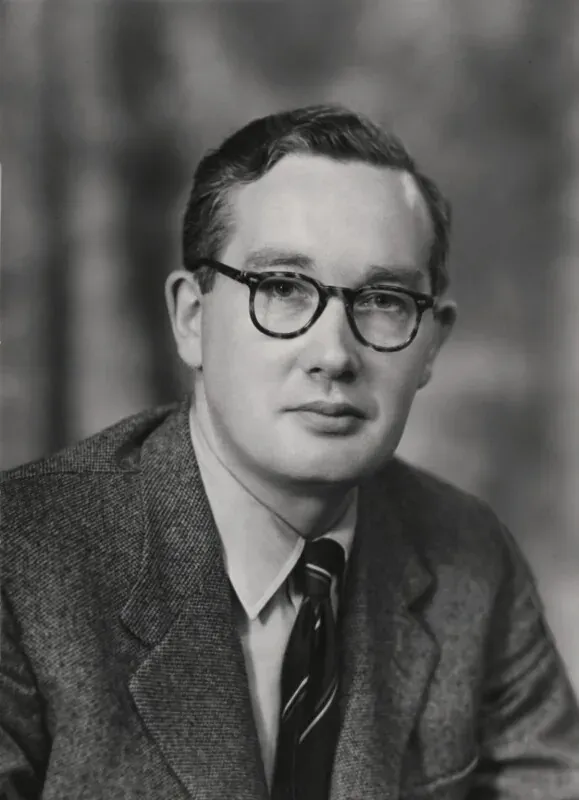 A formal head-and-shoulders photograph of a young man with side-parted hair and dark-framed glasses, wearing a tweed jacket and striped tie.