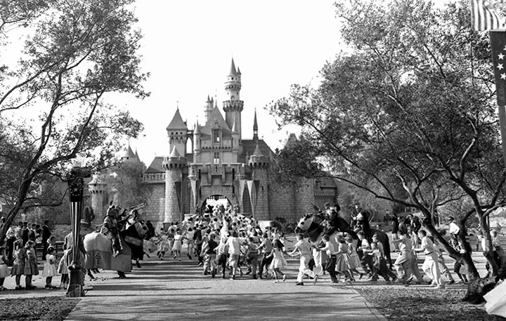 Children running along a tree-lined walkway toward a fairy-tale castle with turrets and a drawbridge, past costumed performers on horseback.