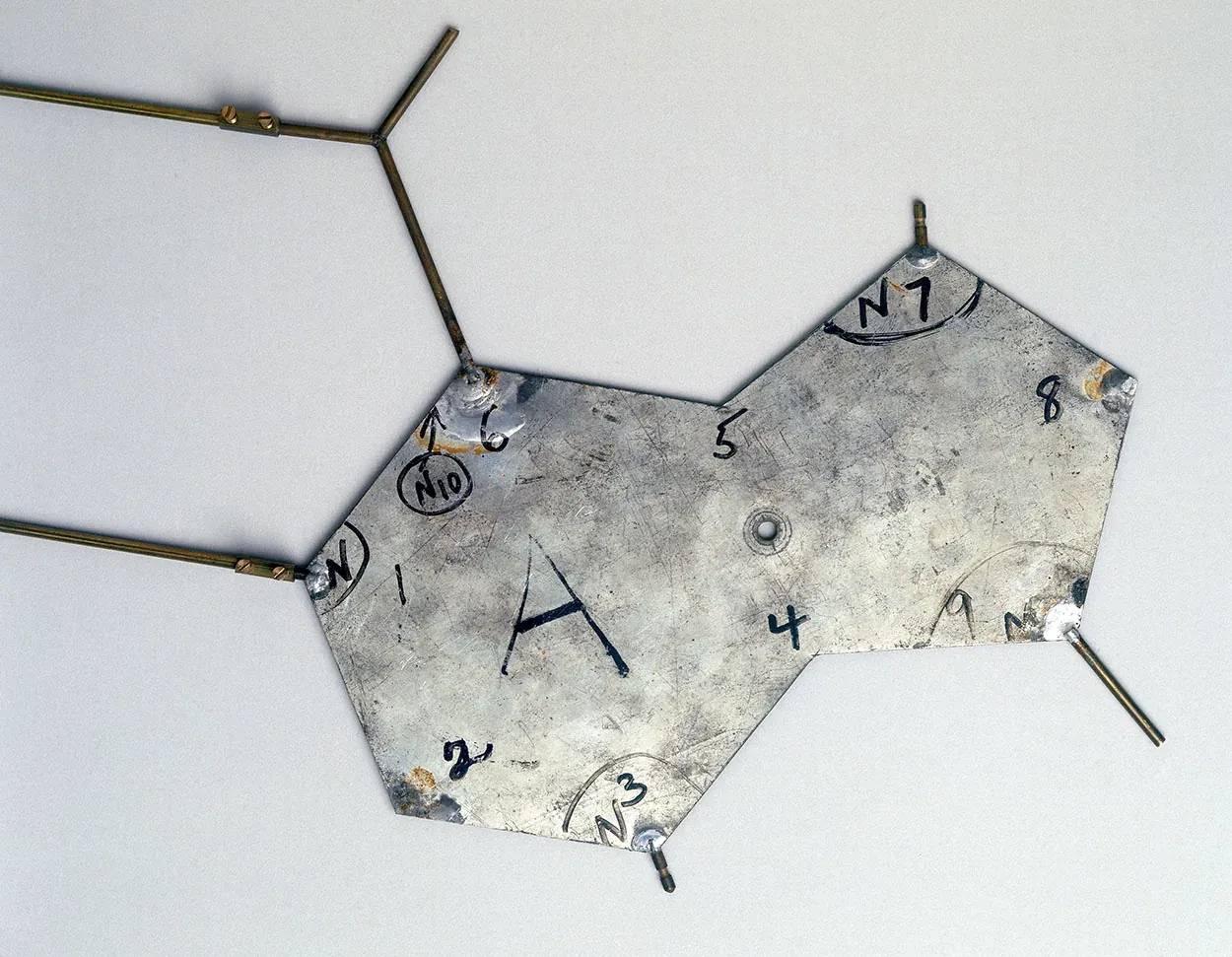 A single flat aluminium plate stamped with the letter A and hand-written atom-numbering marks, with two thin brass rods projecting from one edge, photographed against a plain background.