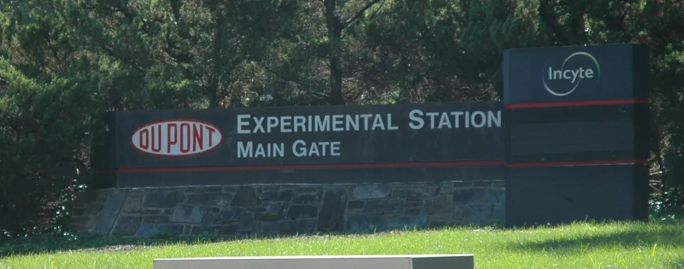 A low stone-and-metal sign reading DUPONT EXPERIMENTAL STATION MAIN GATE, set against a backdrop of trees, with a second sign for a later occupant, Incyte, beside it.