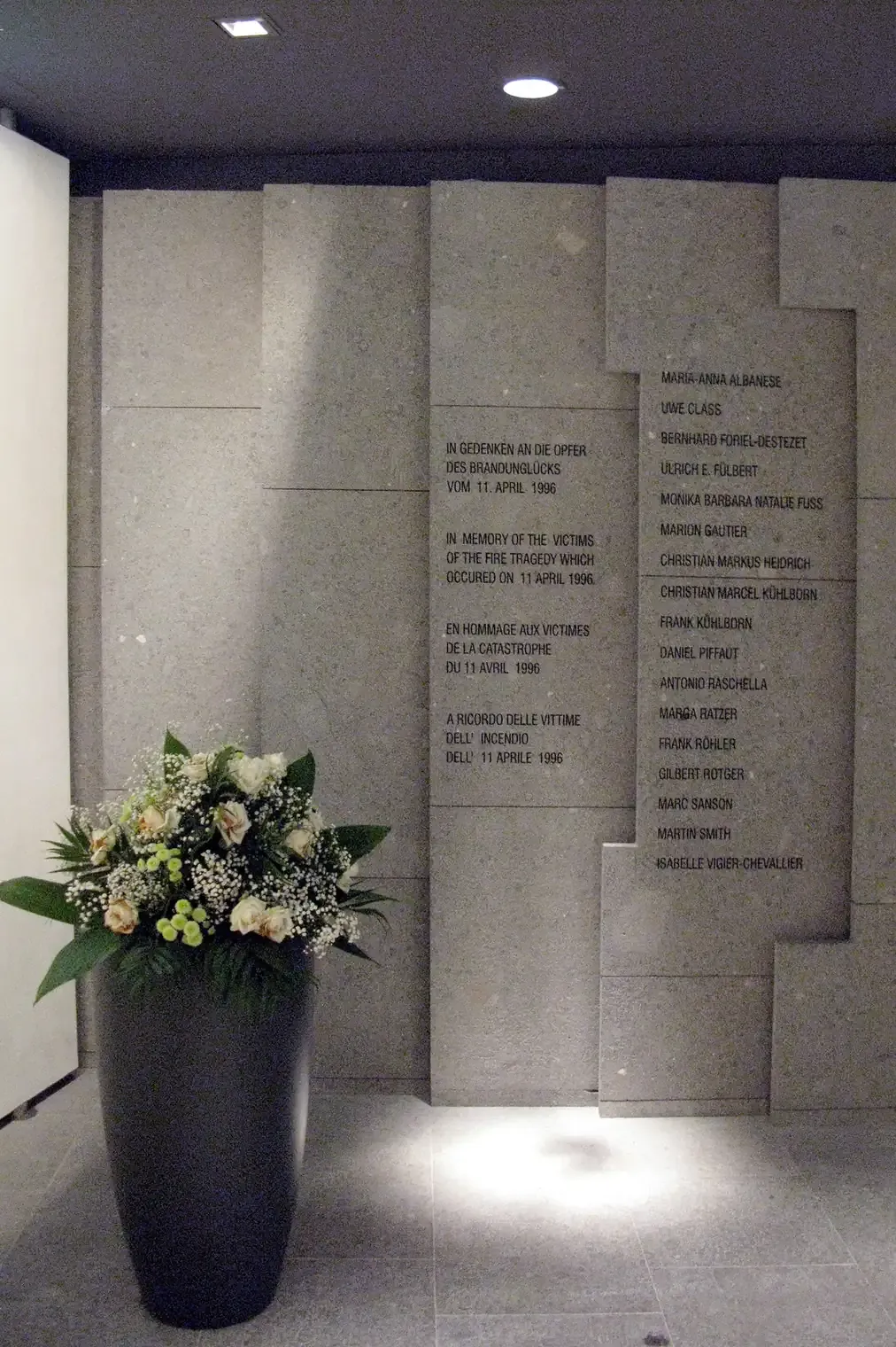 A polished stone memorial wall inside an airport terminal, inscribed in four languages with a dedication to the victims of an April 1996 fire, a column of names beside it, and a tall vase of white flowers standing in front.