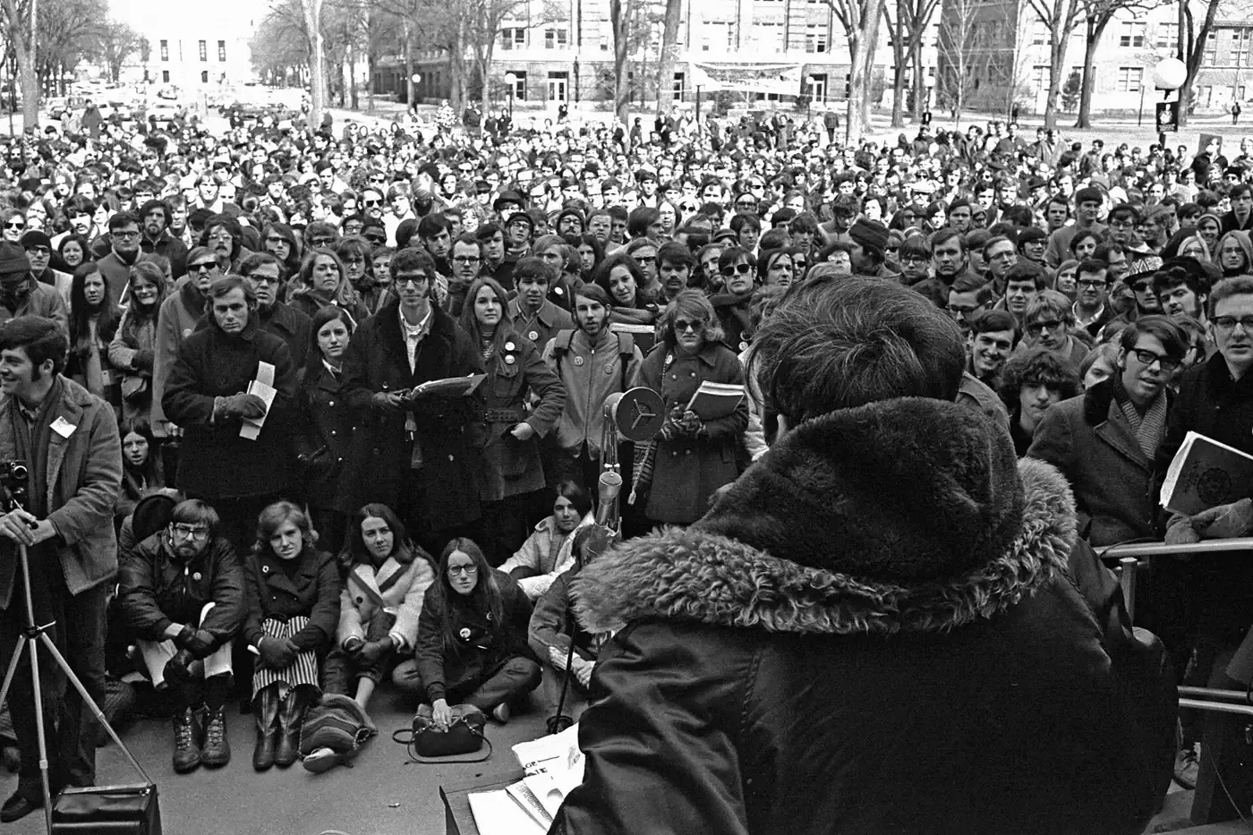 A large crowd of young people, many in winter coats, gathered outdoors around a speaker at a podium with a microphone, seen from behind the speaker.