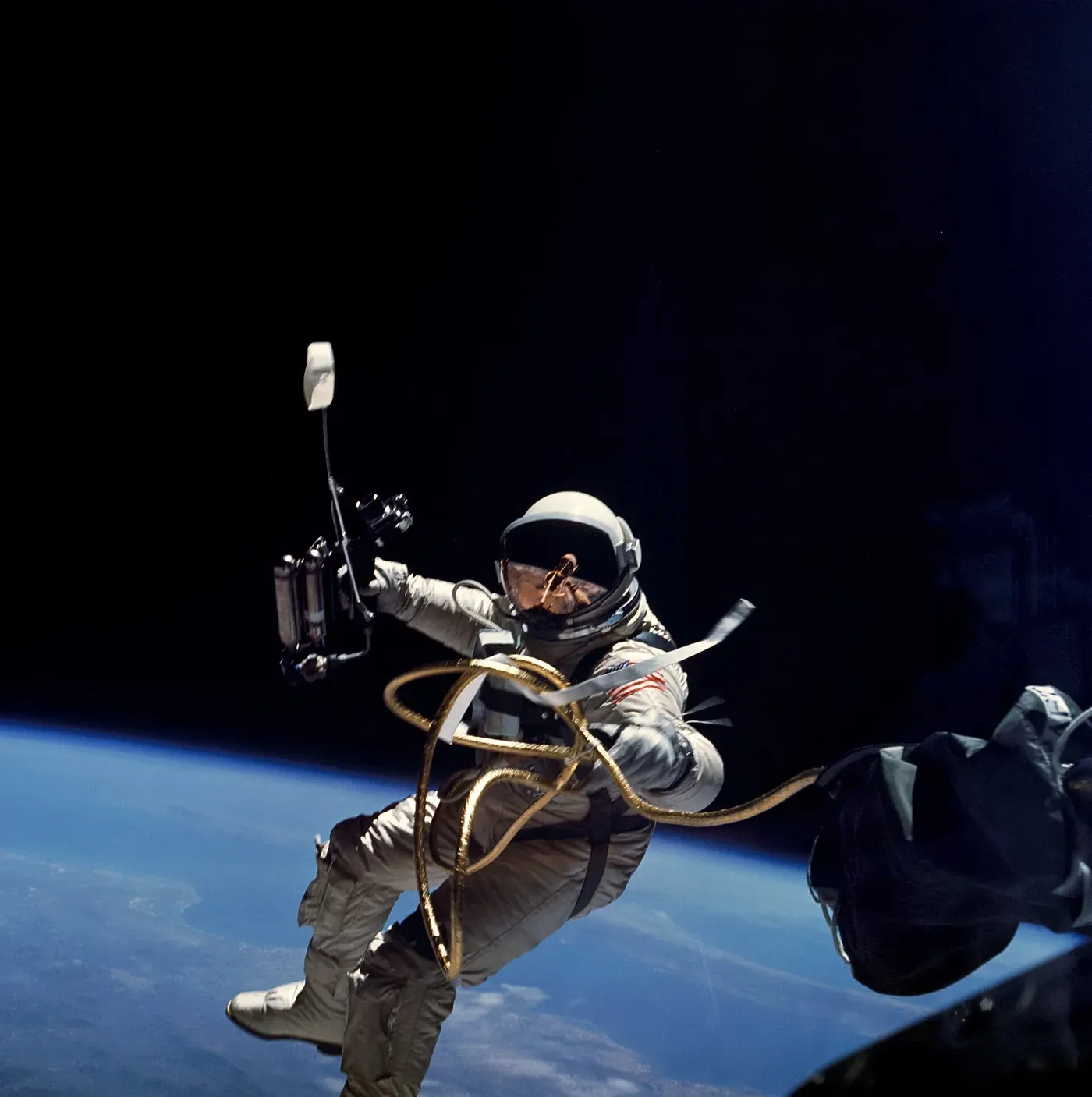 An astronaut in a white spacesuit floating in space above the curve of the blue Earth, tethered by a gold-coloured umbilical line, holding a hand-held manoeuvring unit.