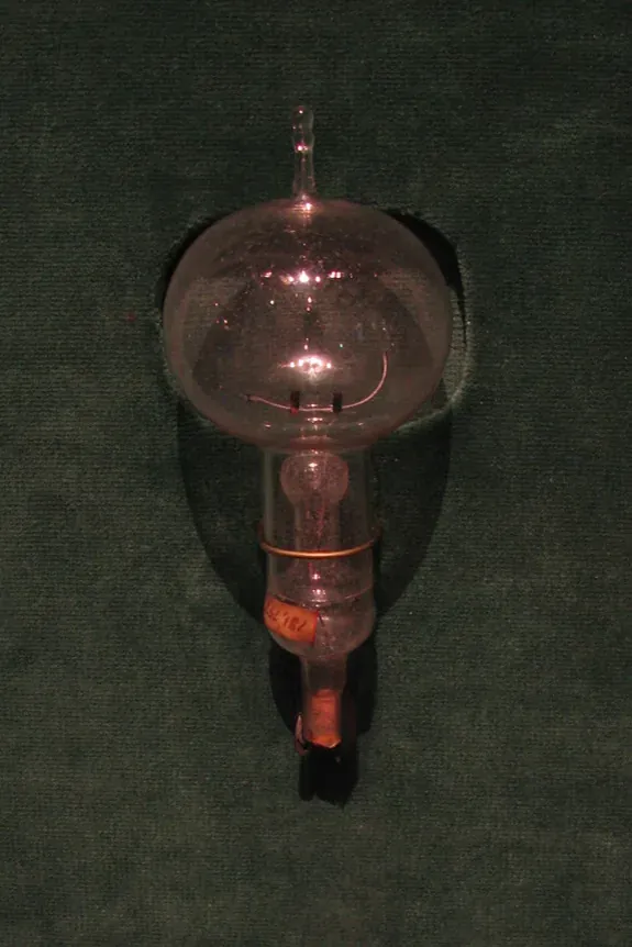 An early Edison incandescent light bulb with a pear-shaped glass envelope, a looped carbon filament, and a screw-style base, mounted against a dark green background.