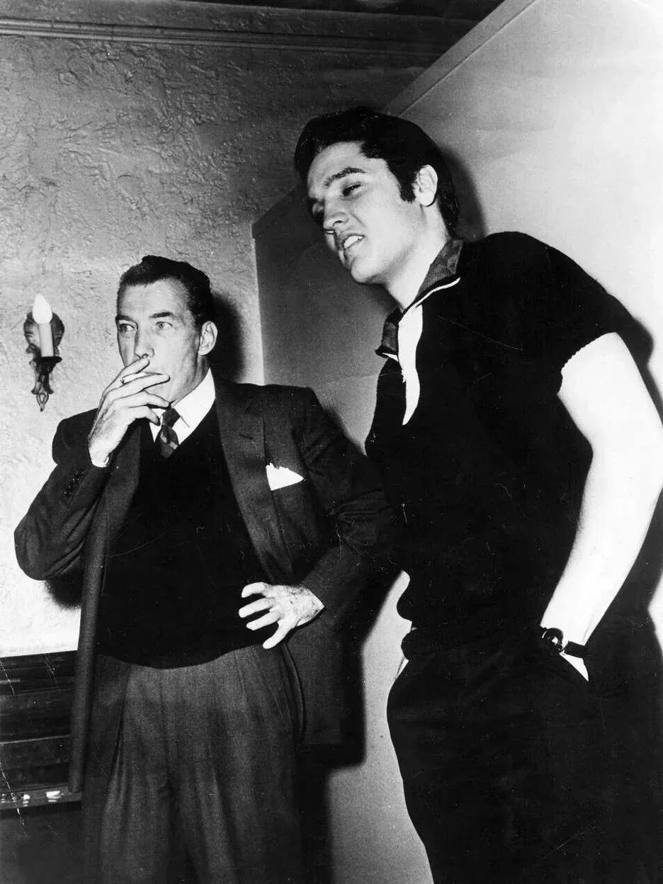 Black-and-white photograph of Elvis Presley, in a dark shirt with an open collar, standing beside Ed Sullivan, who holds a lit cigarette, backstage.