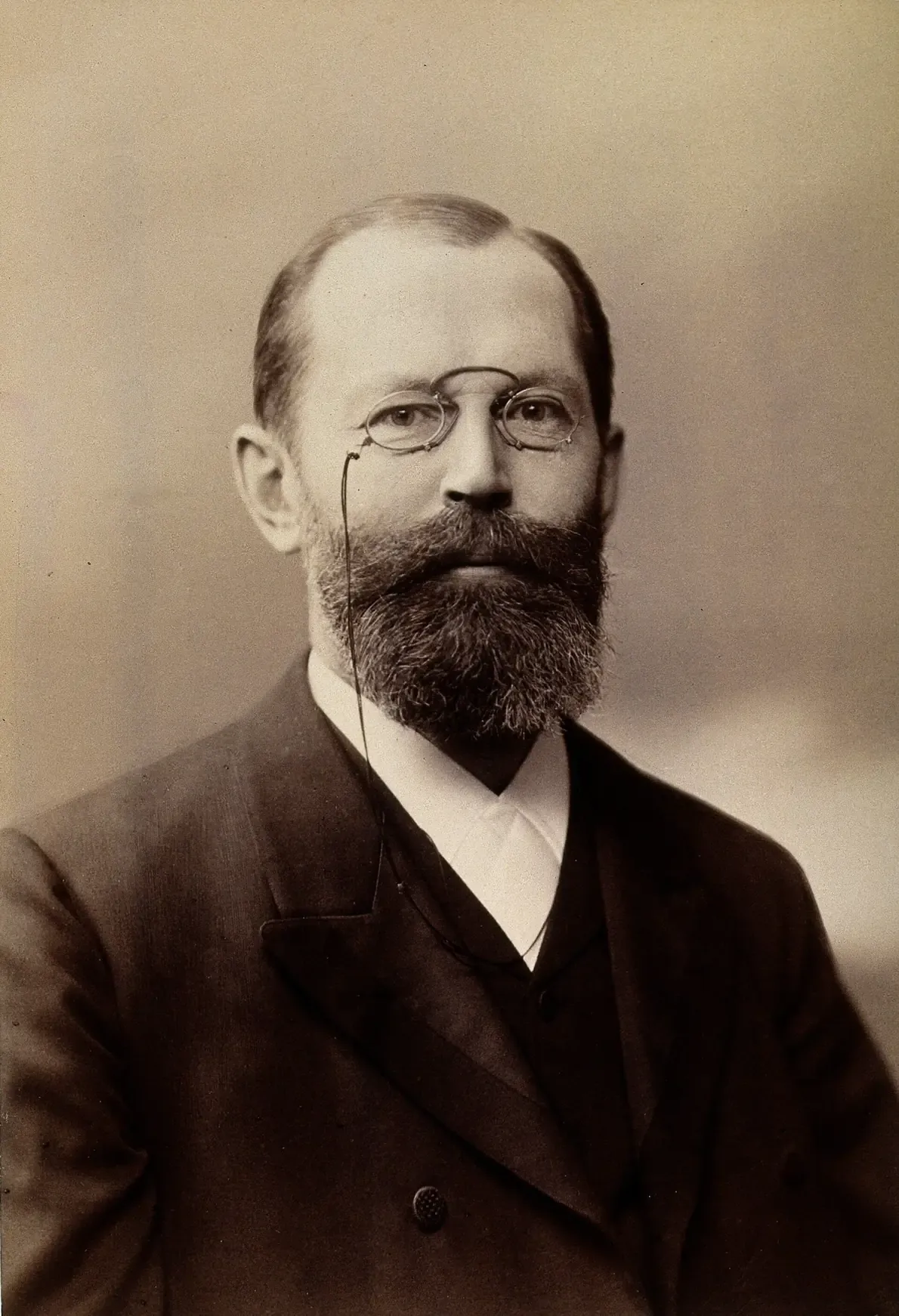 A formal studio photograph of a bearded man with wire-rimmed pince-nez glasses and a receding hairline, wearing a dark suit and high collar.