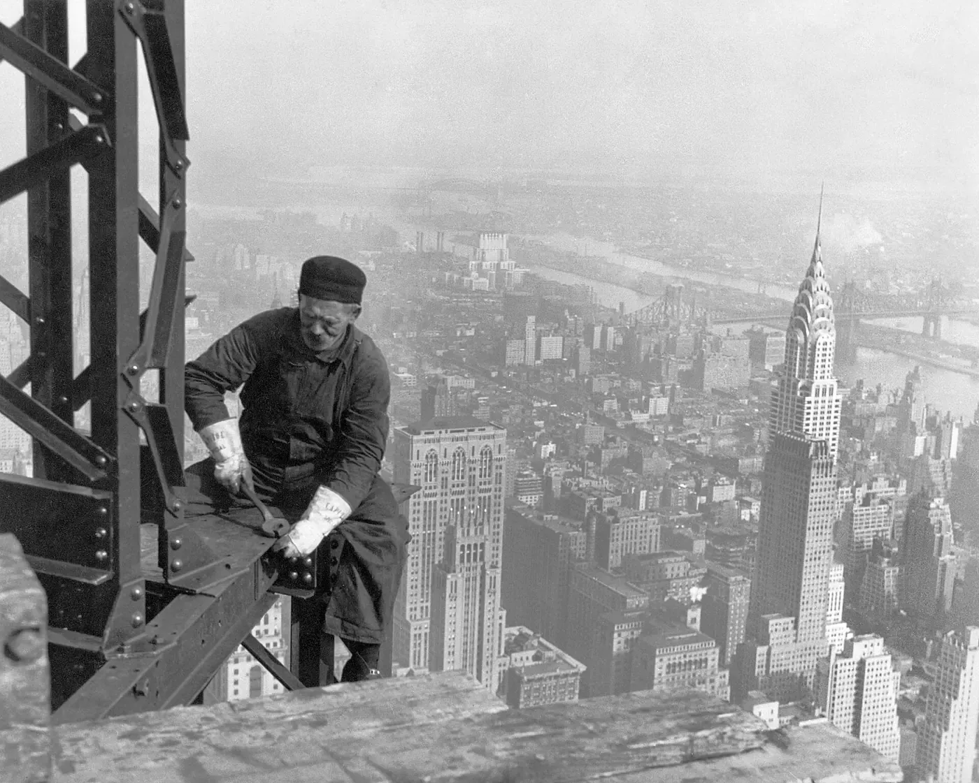 A steelworker crouches on a girder high above Manhattan, hammering a rivet, with the Chrysler Building and the East River visible far below in the haze.