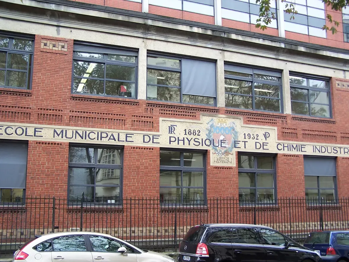 A red-brick industrial-style school building with large multi-pane windows, a stone plaque reading 'École Municipale de Physique et de Chimie Industrielles' set between dates 1882 and 1932, and cars parked along the street.
