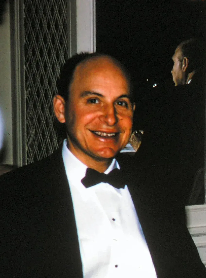 A balding man in a black bow tie and tuxedo, smiling broadly at the camera at an evening event, with another figure visible in a dark doorway behind him.