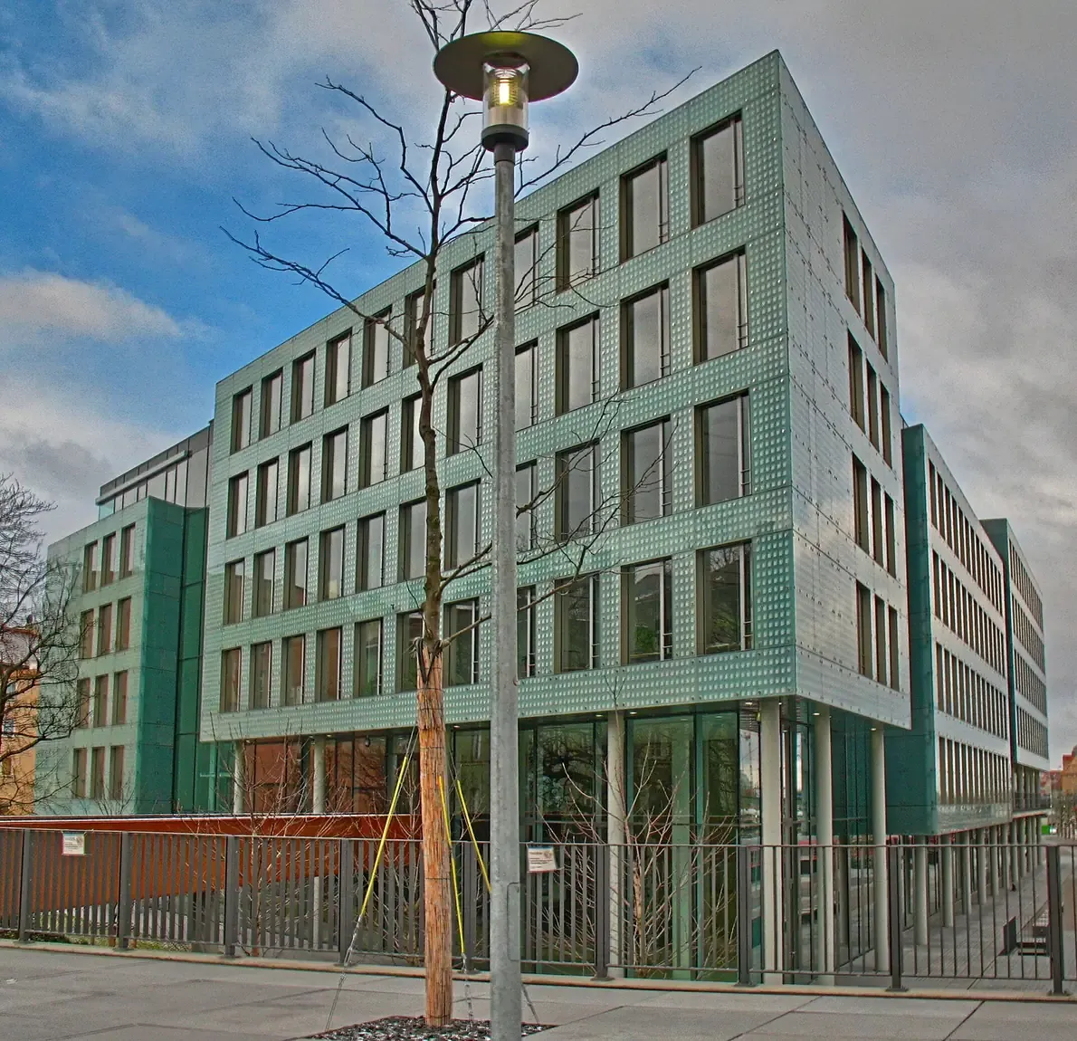 A modern multi-storey office building clad in perforated green metal panels, photographed from a street corner with a lamp post and bare young trees in front.