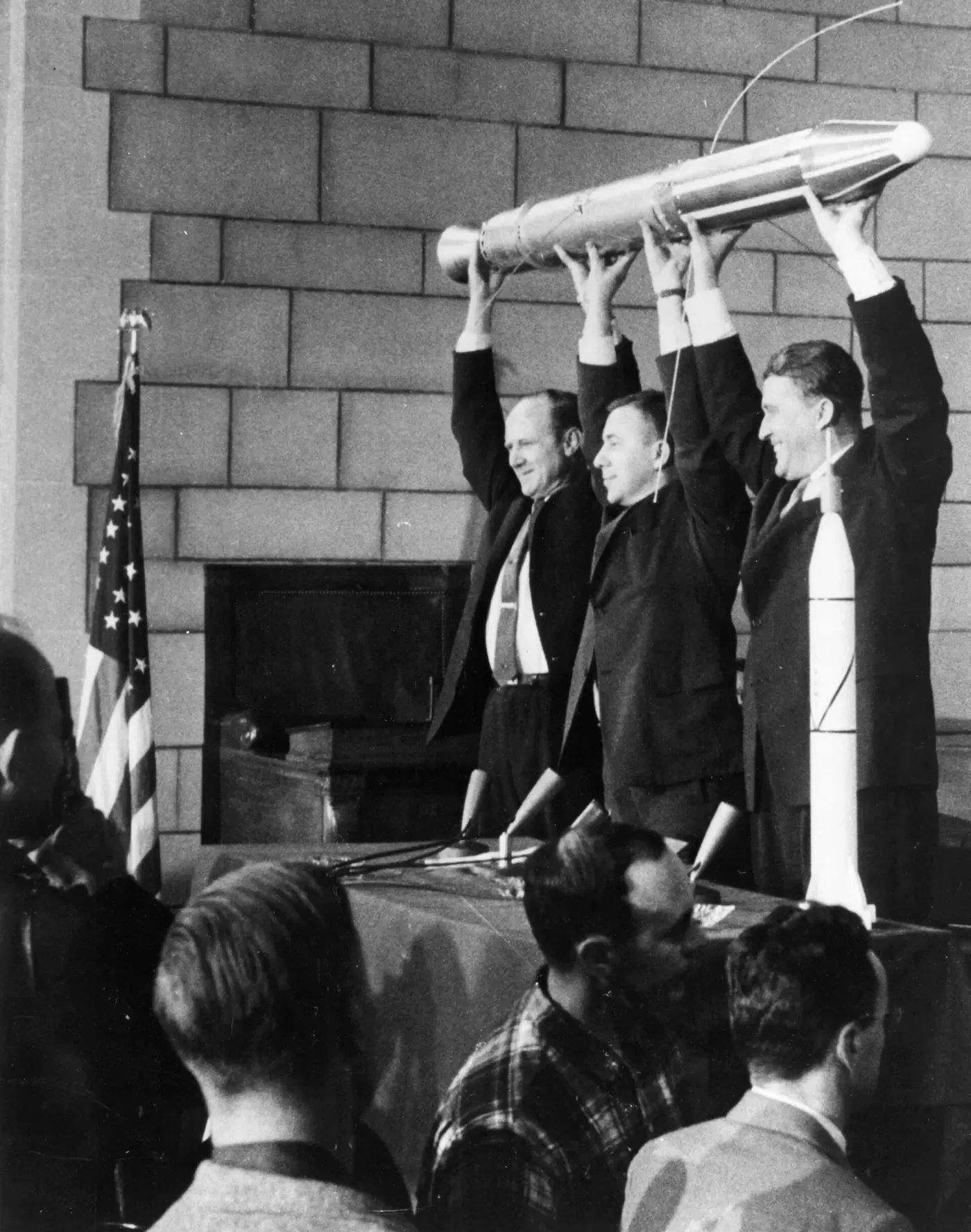 Three men in suits stand on a stage before an audience and a microphone, holding a long, slender satellite model above their heads with both hands, in front of a stone wall and an American flag.