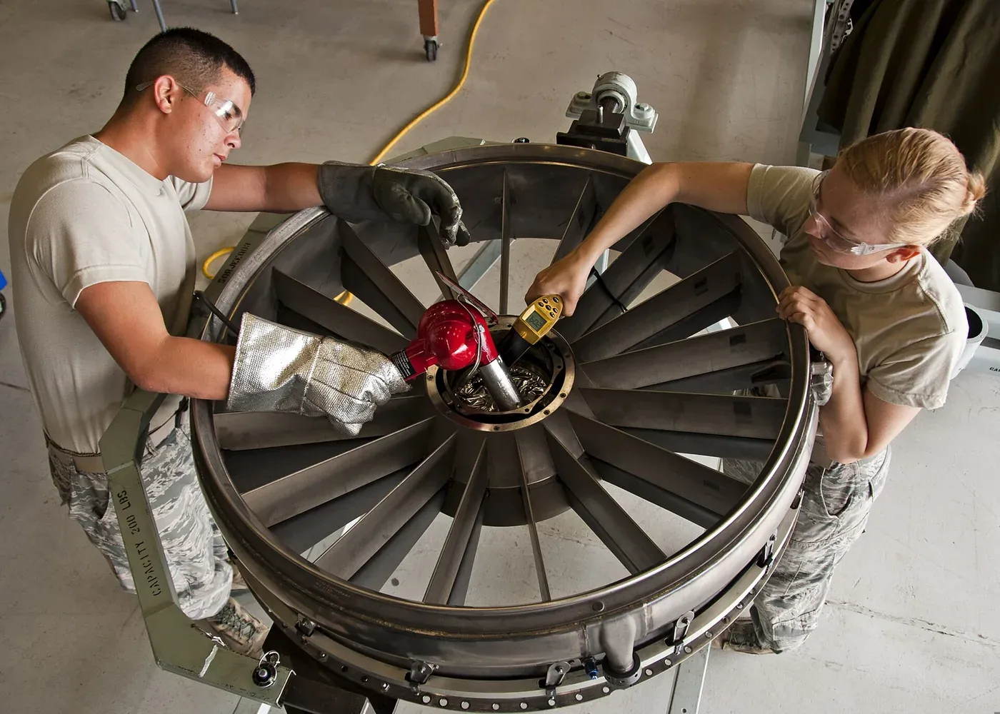 Two US Air Force trainees in tan uniforms work on a large turbine bearing housing during aircraft engine maintenance.