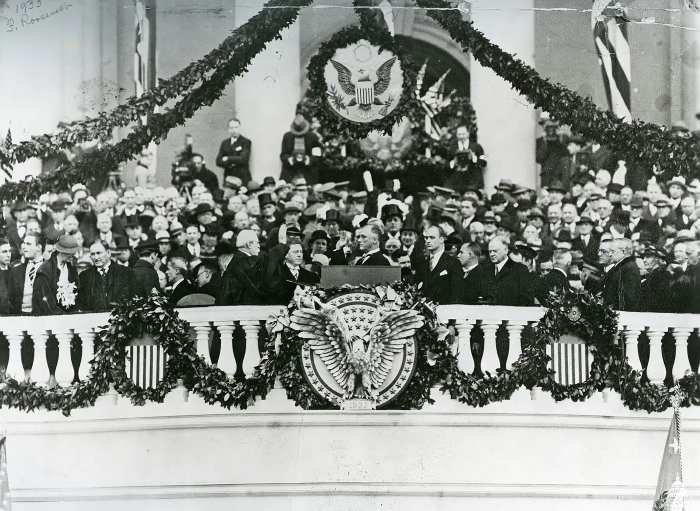 A crowd-filled inaugural platform draped in garlands, where a man in a dark coat raises his right hand to take an oath before a robed official, flanked by dignitaries in overcoats and top hats, with the year 1933 carved into the balustrade below.