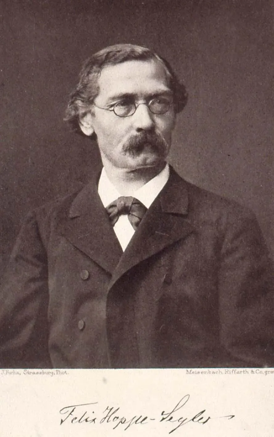 A sepia-toned 19th-century portrait of a man with round spectacles, a moustache, and a patterned bow tie, with his signature printed below the image.