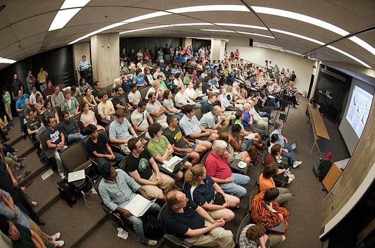An overhead, fisheye view of a packed university auditorium, with more than two hundred people seated and standing to watch a projection screen at the front displaying a scientific slide.