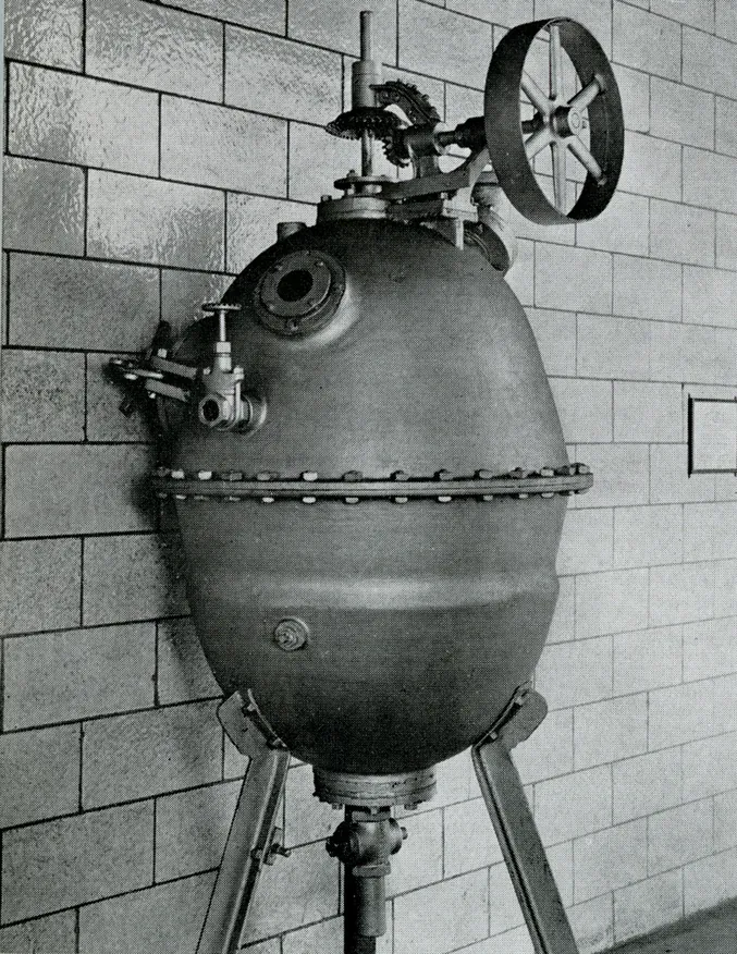 An egg-shaped riveted iron pressure vessel standing on a tiled laboratory floor, with a large valve wheel on top and pipework running to the wall.