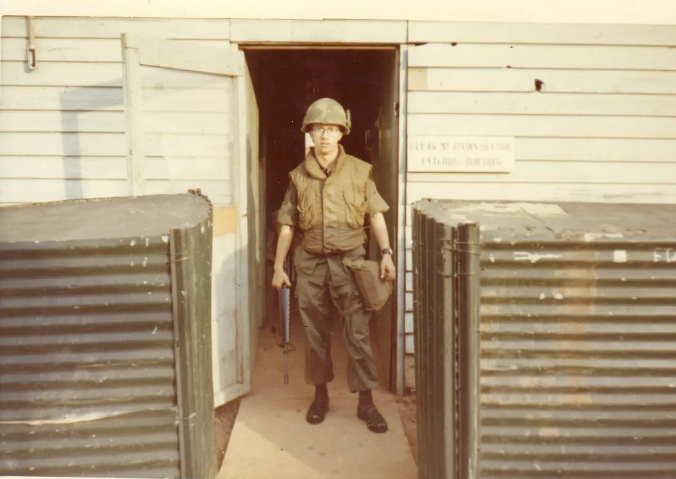 A young U.S. Army soldier stands in a doorway between head-high stacks of corrugated metal sheeting, wearing a bulky fabric flak vest over his fatigues and holding a shotgun, Vietnam, 1971.