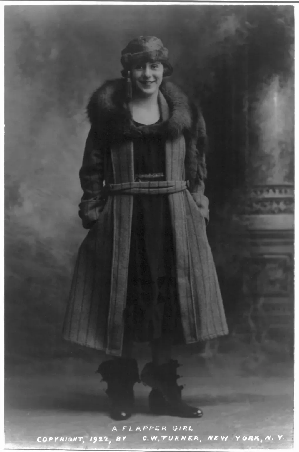 A young woman in a fur-collared tweed coat and cloche hat, hands in her pockets, posing for a studio photograph captioned 'A Flapper Girl'.