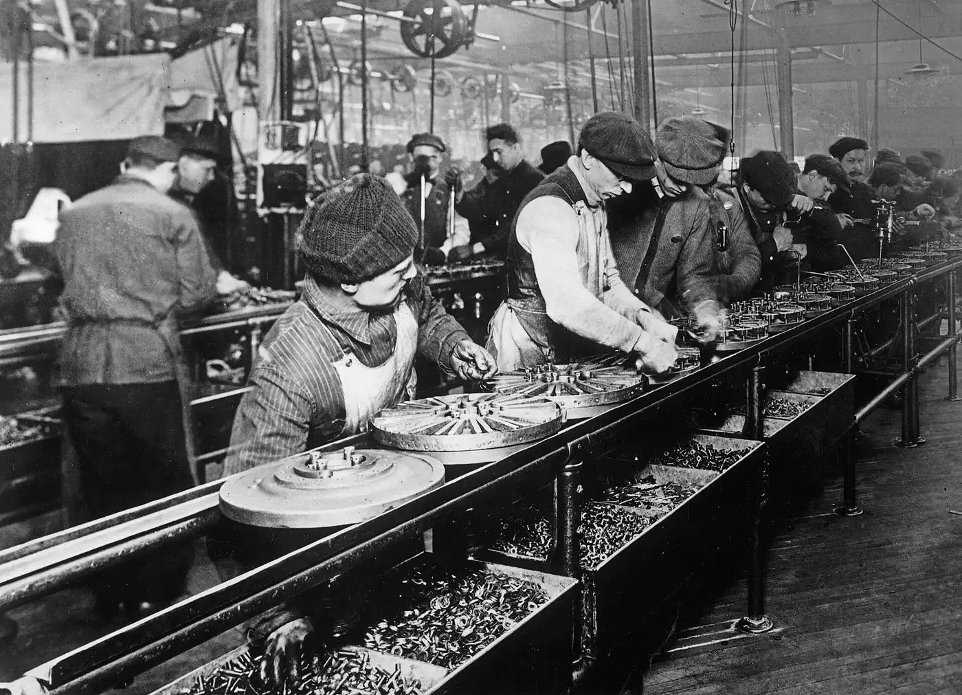 Black-and-white photograph of workers in caps and overalls assembling wheel hubs and small metal parts along a moving conveyor line, with bins of components at their feet.