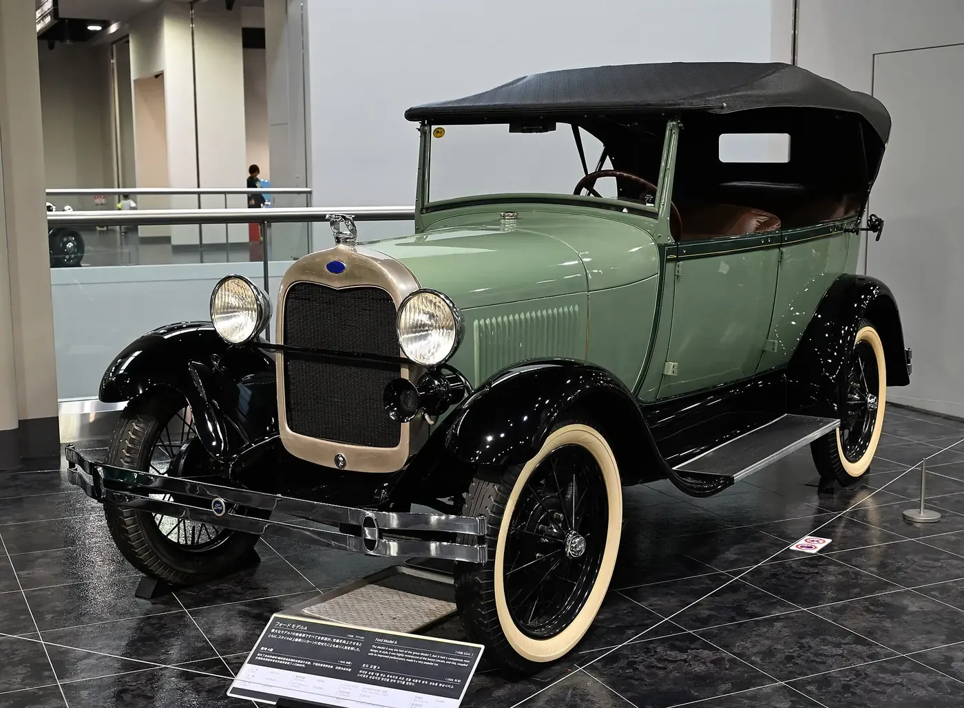 A restored 1928 Ford Model A touring car in pale green and black, with a folded black canvas top, on display in a museum with an informational placard in front of it.