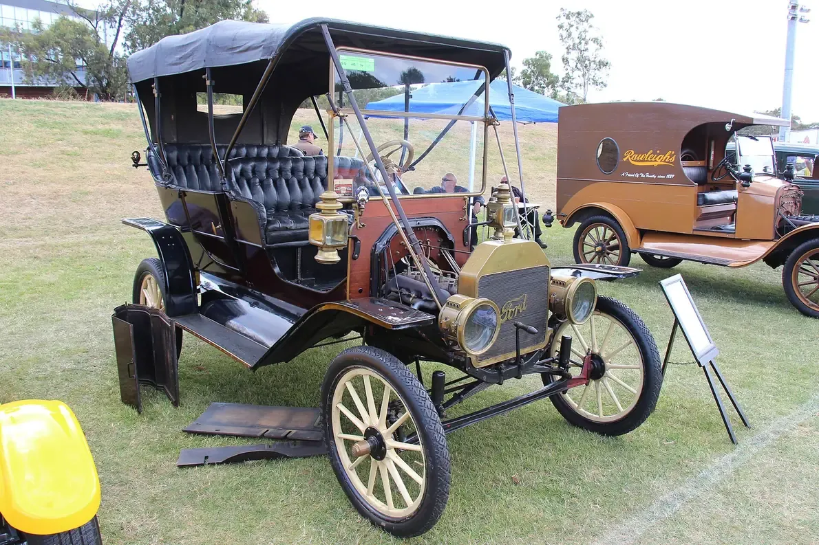A restored early touring car with a folding canvas roof, brass headlamps and wooden-spoked wheels, parked on grass at a vintage vehicle show.