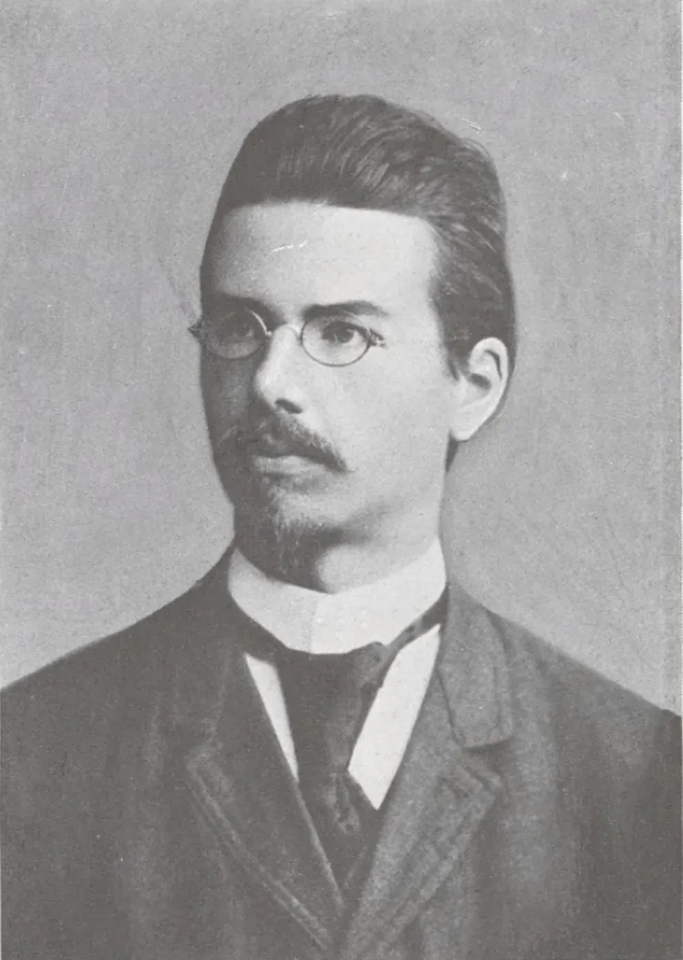 A formal black-and-white studio portrait of a young man with round wire-rimmed glasses, a moustache and small beard, wearing a high-collared shirt, tie and dark jacket.